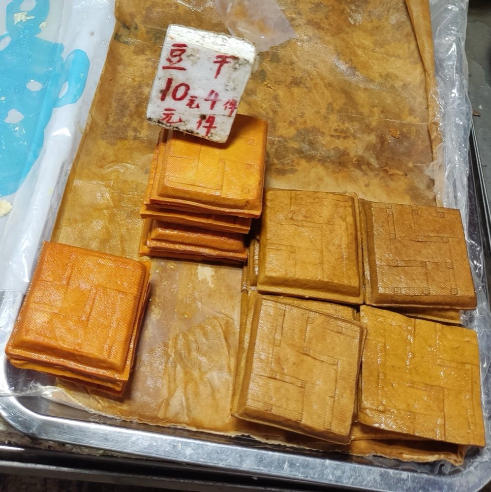 市场摊上一盘金棕色的豆干方块，旁边立着手写的「豆干」价格牌